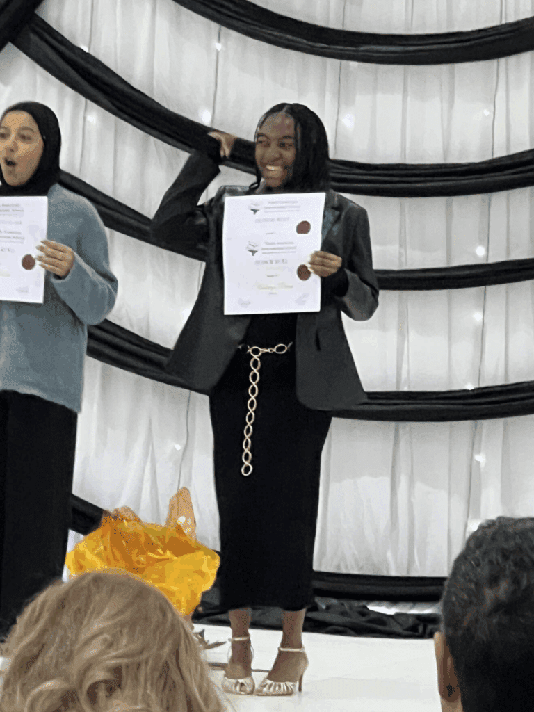 Two women on stage proudly holding up papers, smiling at the audience during a presentation.