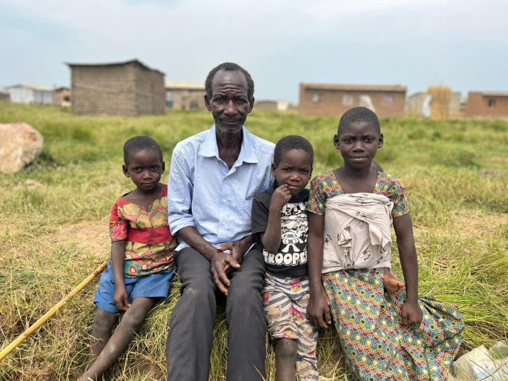 An elderly man sits on grass with three children, two girls and a boy, in a rural setting. They wear colorful, traditional clothing, against a backdrop of simple homes.